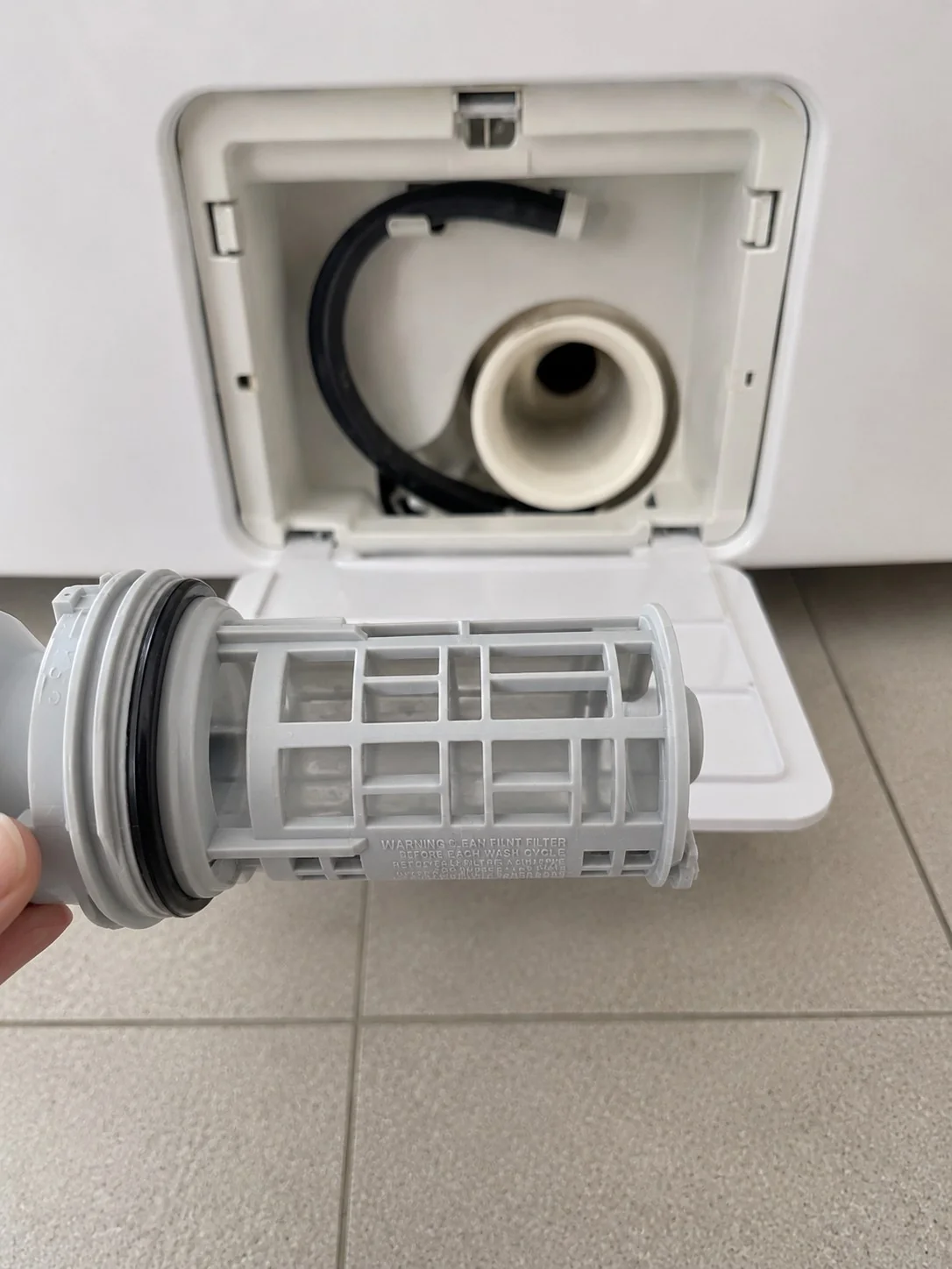 The same drain pump filter cartridge after cleaning, grey plastic mesh clear of debris in front of the same access panel